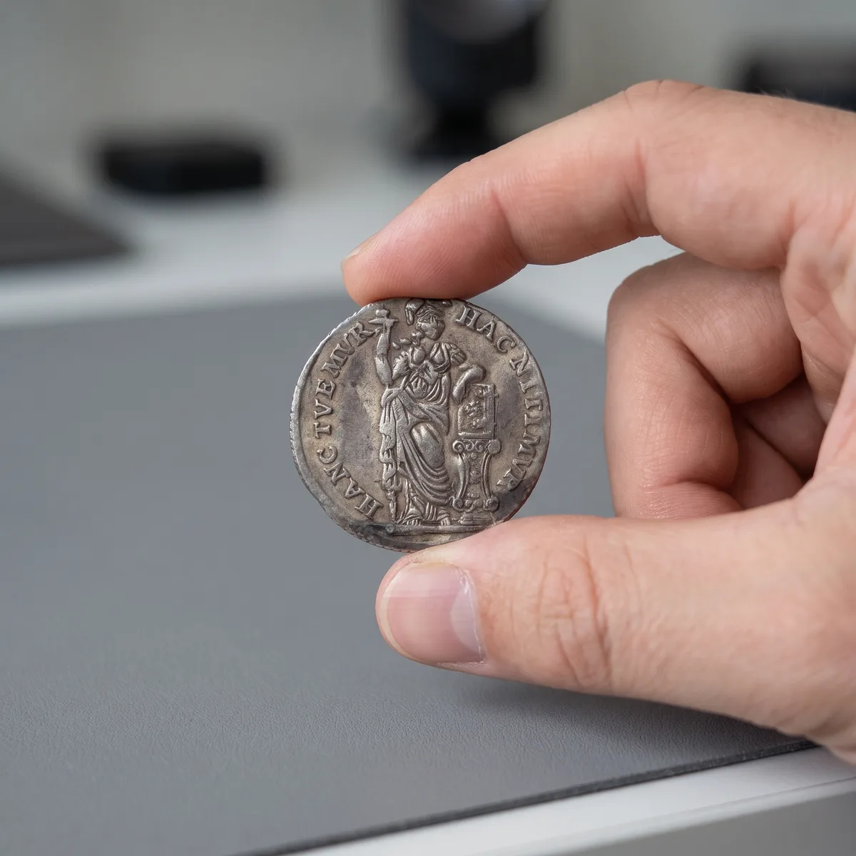 Twee vingers houden een zilveren VOC-gulden voorzichtig bij de rand vast, boven een grijze werkmat op een bureau